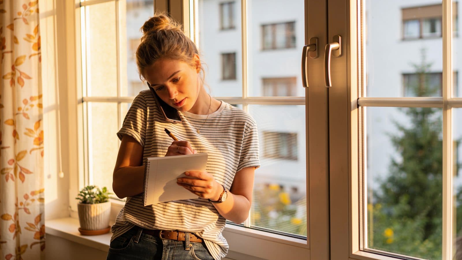 Junge Frau telefoniert am Fenster mit dem Handy am Ohr und macht sich dabei eine Notiz