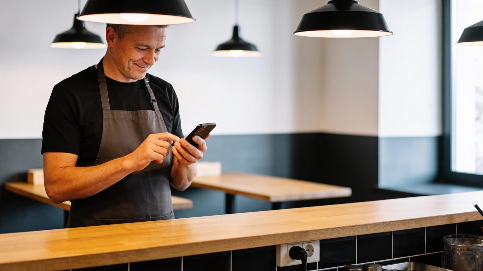 Gastronom in Schürze steht hinter seinem Tresen und schaut interessiert ein kurzes Video auf seinem Smartphone an